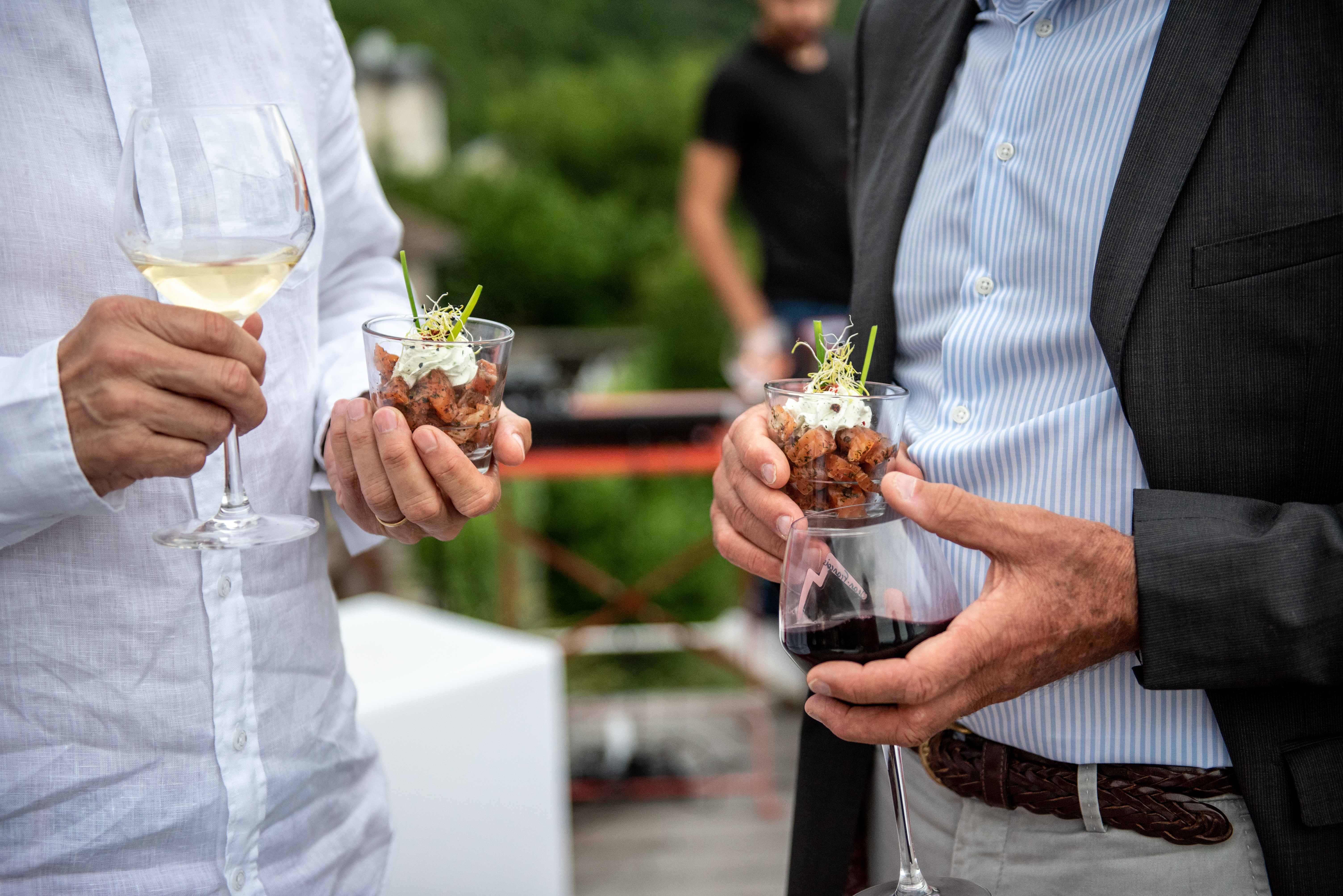 Gros plan sur deux invités lors d'un cocktail en extérieur, chacun tenant une verrine gastronomique et un verre de vin, avec la végétation en arrière-plan.