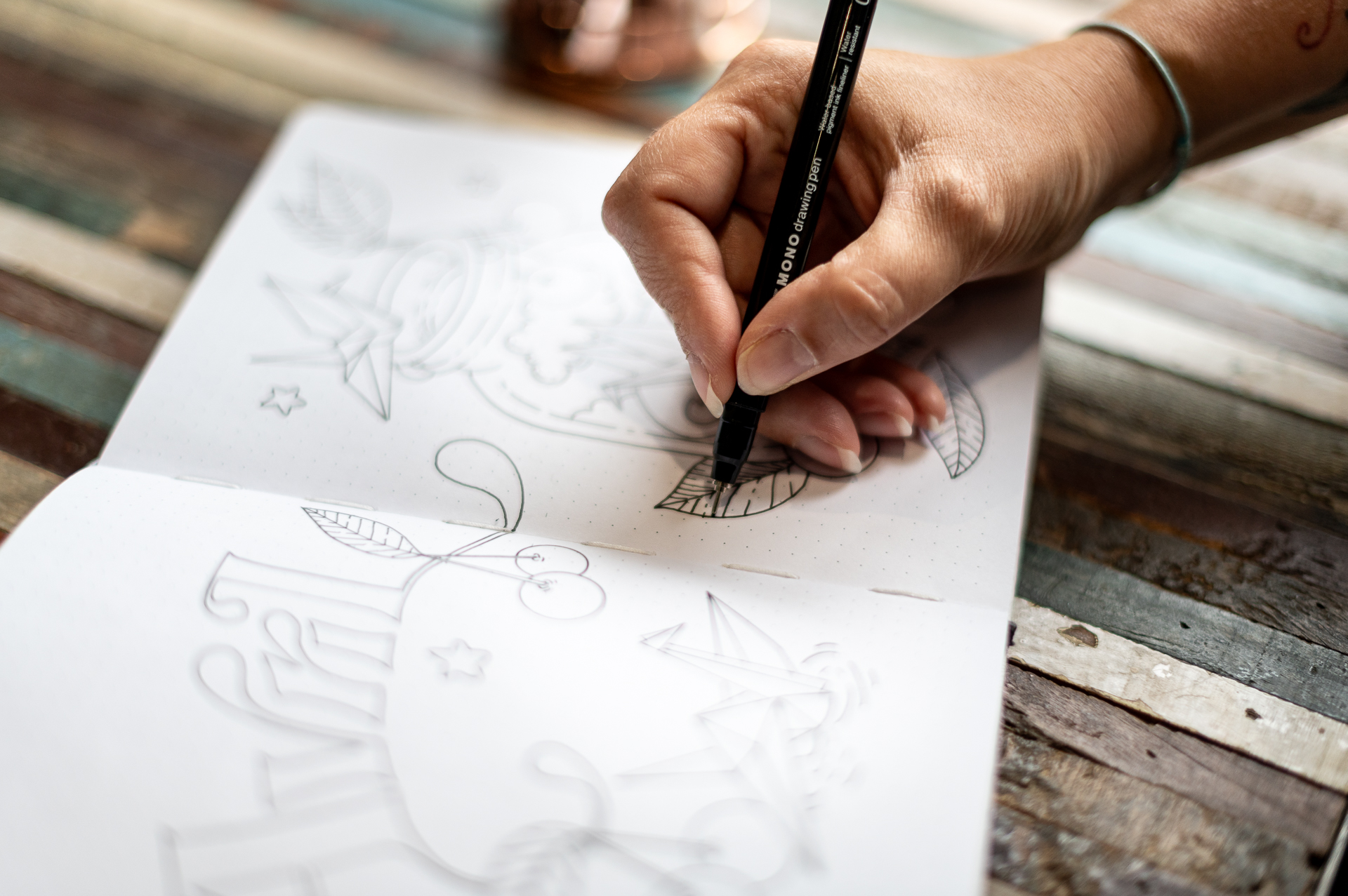 Gros plan sur une main dessinant au crayon noir des esquisses de motifs floraux et de lettrages dans un carnet, pour la marque Enemesis and Art.