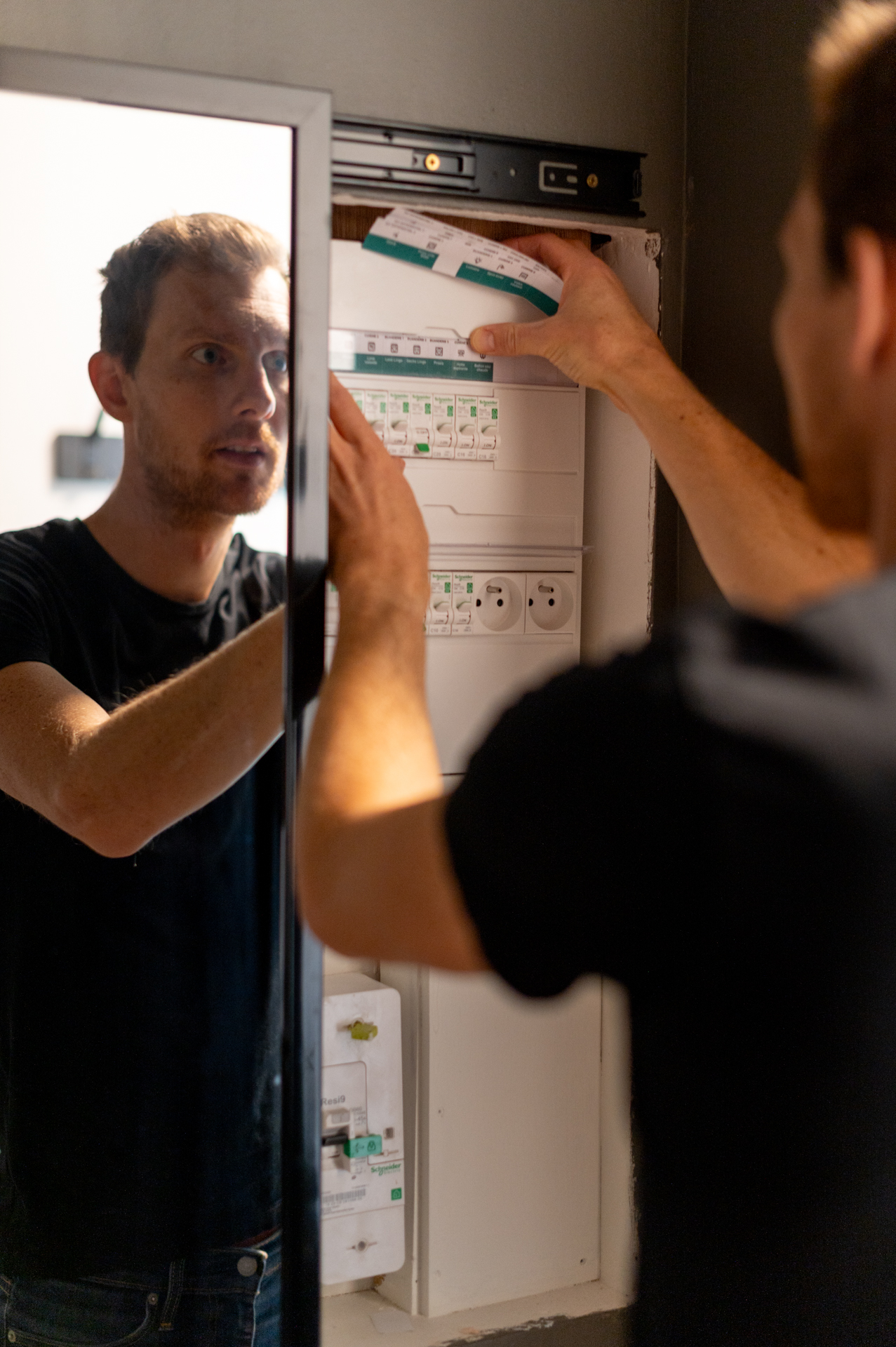 Un électricien de TP Elec intervient sur un tableau électrique ouvert, main levée vers les disjoncteurs, avec son reflet visible dans un miroir à côté.