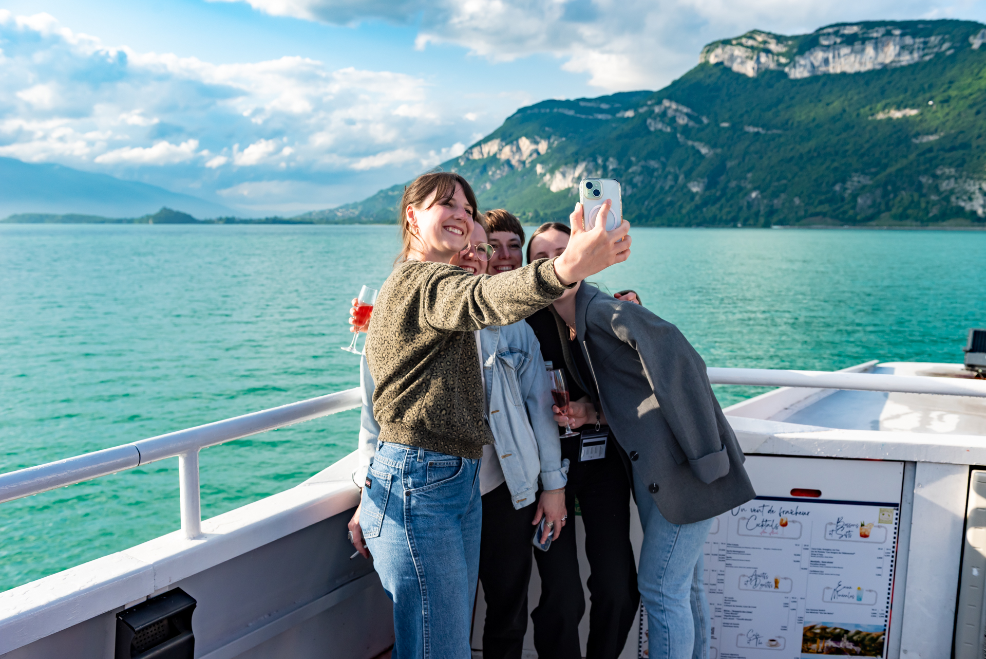 Groupe de personnes prenant un selfie au smartphone, un verre à la main, à bord d'un bateau sur le lac du Bourget, avec la montagne et l'eau turquoise en arrière-plan.