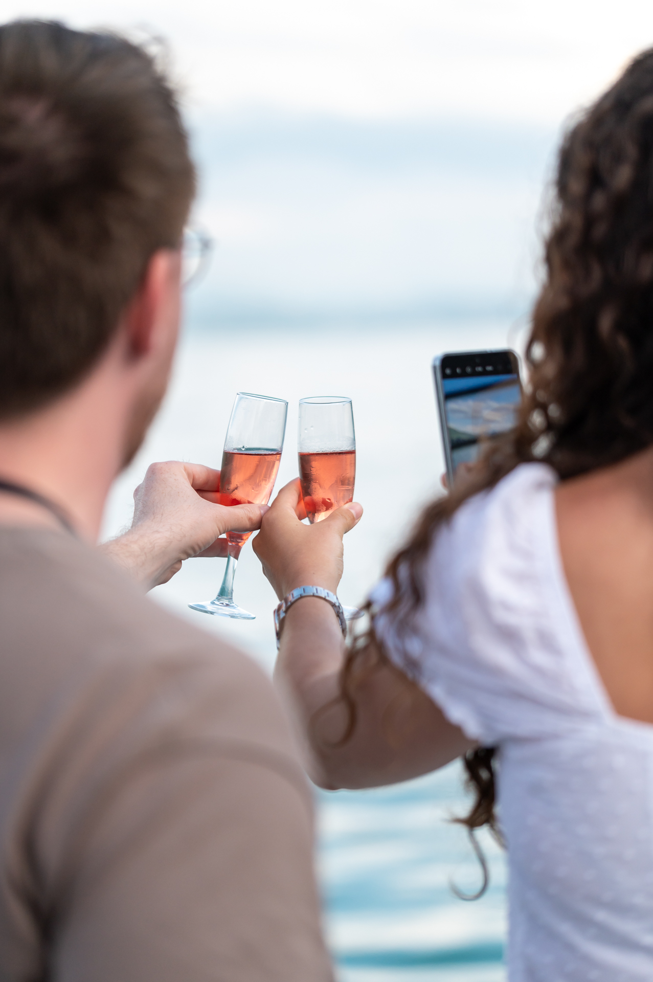 Deux personnes de dos trinquent avec des verres de vin rosé au bord du lac du Bourget, l'eau et les reflets clairs en arrière-plan.