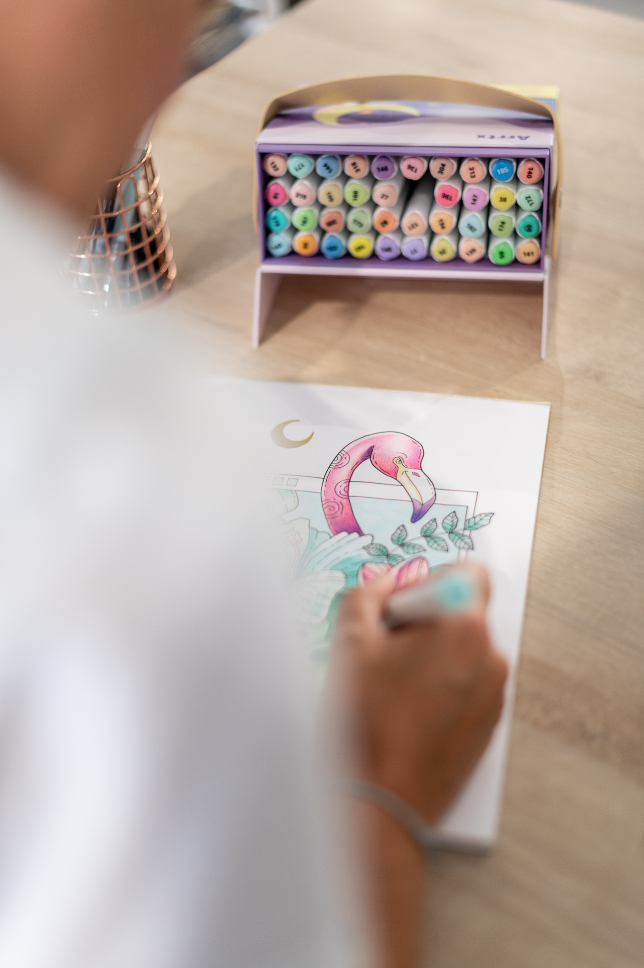 Gros plan sur les mains d'une graphiste en train de dessiner un flamant rose aux feutres de couleur, pour la marque Enemesis and Art, avec un porte-feutres coloré en arrière-plan.