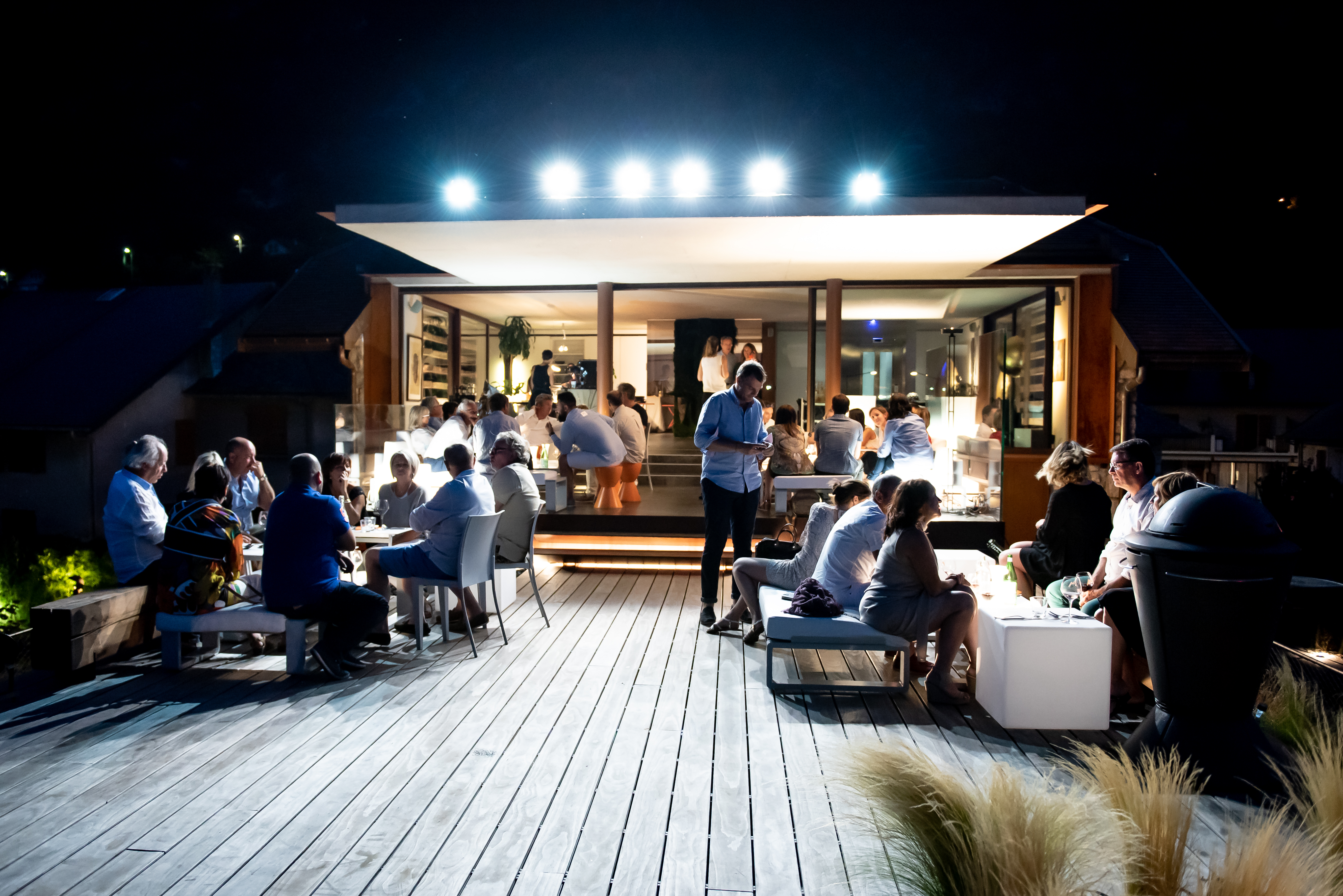 Terrasse en bois du JMV Resort illuminée à la tombée de la nuit lors de la soirée d'inauguration, les invités installés en petits groupes autour de la structure éclairée au bord du lac du Bourget.