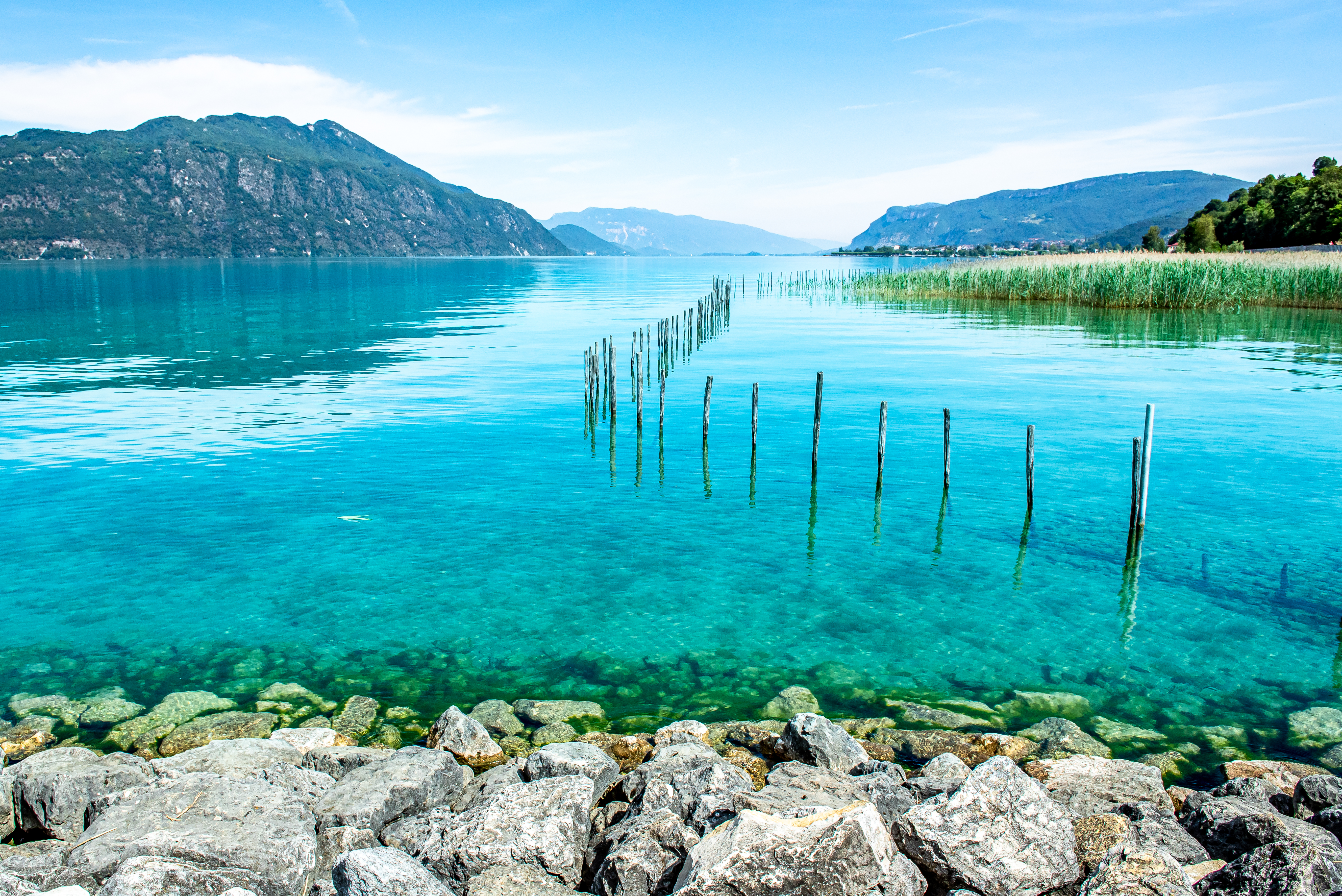 Vue sur les eaux turquoise et transparentes du lac du Bourget depuis la rive, avec des rochers au premier plan et d'anciens pieux en bois plantés dans l'eau, montagnes en arrière-plan.