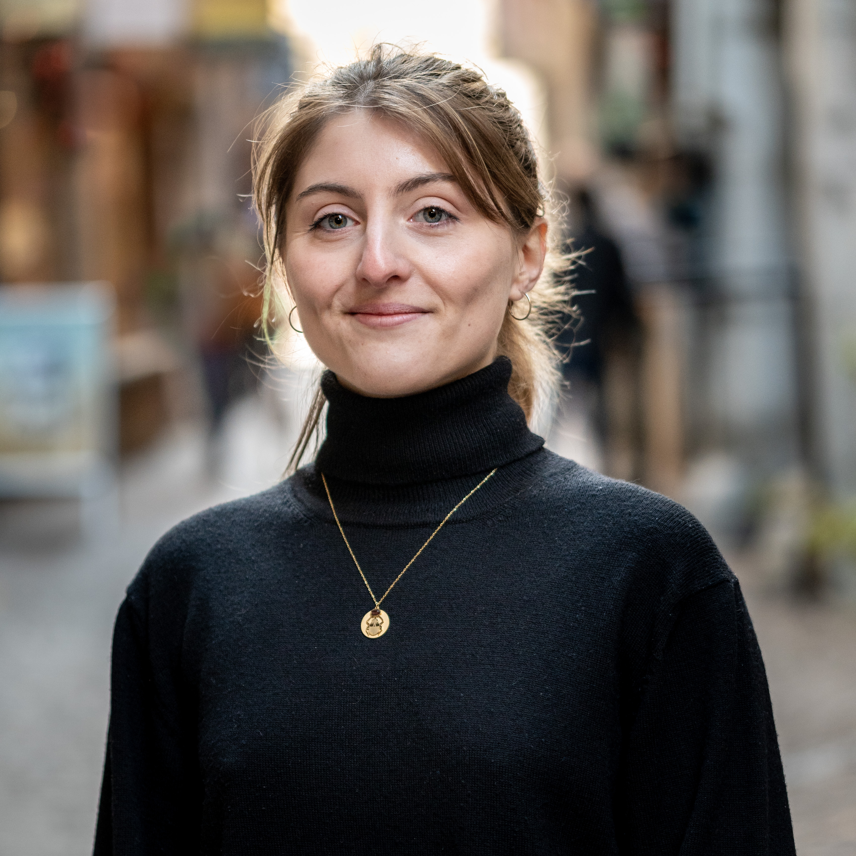Portrait d'une femme aux cheveux tressés, col roulé noir et collier doré, prise en extérieur dans une rue animée avec un arrière-plan flouté.