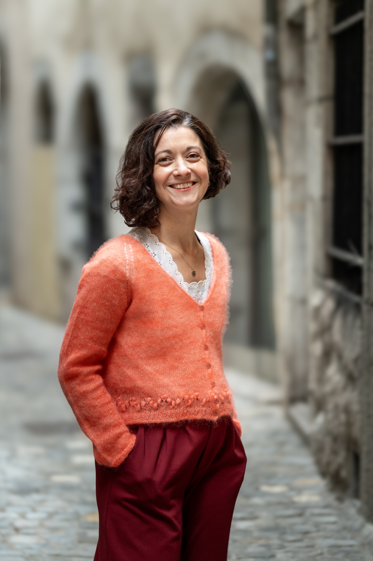 Portrait en pied de Céline Messina, fondatrice de Mlle Pétronille, gilet orange et pantalon bordeaux, dans une ruelle avec des arches en pierre.