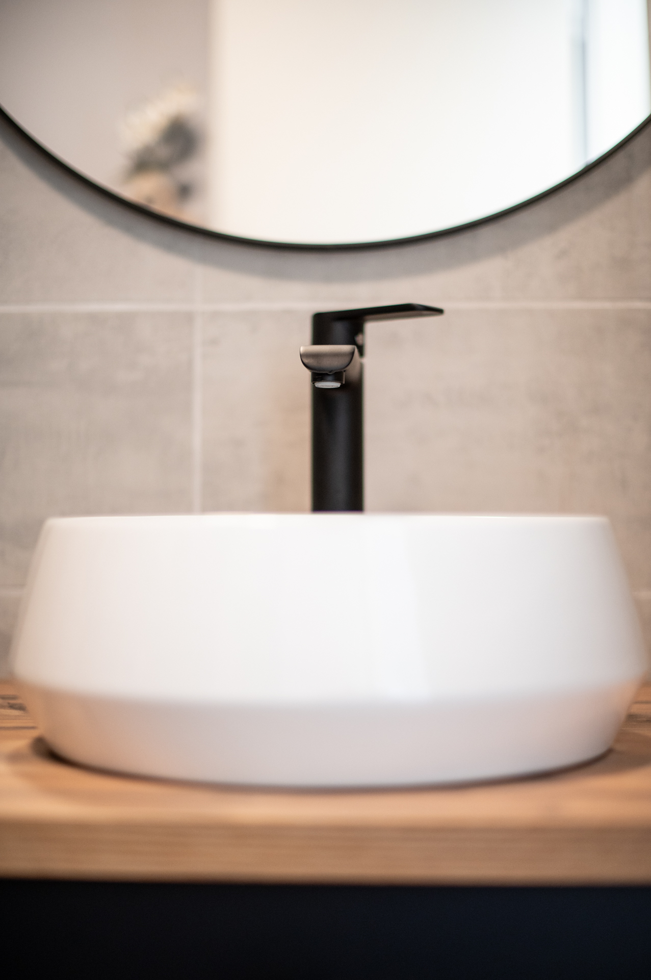 Vasque ronde blanche posée sur un plan de travail en bois, avec une robinetterie noire et un miroir rond au mur, dans une salle de bain moderne aux tons épurés.